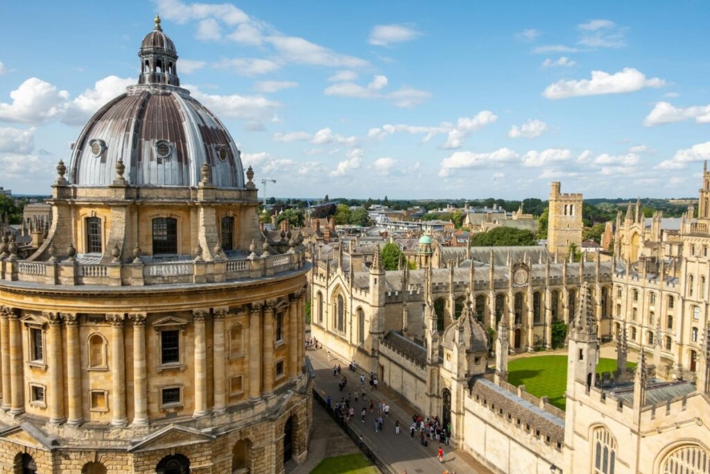 Vista panorâmica da Universidade de Oxford em um dia ensolarado, destacando a arquitetura histórica com cúpulas, torres e prédios medievais que lembram os cenários mágicos de Harry Potter, incluindo pátios acadêmicos e ruas tradicionais da cidade de Oxford, na Inglaterra.