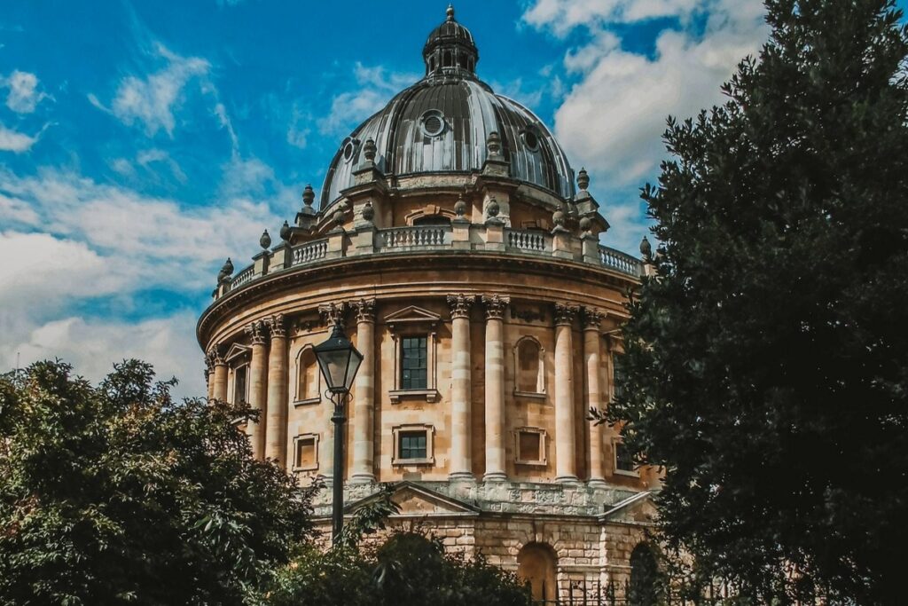 Imagem da Biblioteca Bodleiana Universidade de Oxford, na Inglaterra, destacando a arquitetura neoclássica com grande cúpula histórica, cercada por árvores e céu azul, cenário real do universo mágico de Harry Potter.