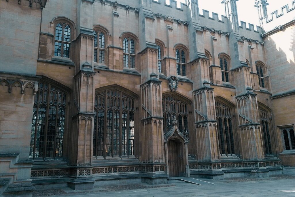A faculdade Divinity School com arquitetura gótica inglesa, destacando grandes janelas arqueadas, detalhes medievais em pedra e entrada ornamentada, cenário real dos filmes Harry Potter.